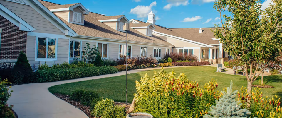 A well-maintained outdoor garden area of a senior living facility with a curved concrete walkway, green grass, various shrubs, flowering plants, and trees. The building has beige siding with white trim and multiple windows, under a blue sky with some clouds.