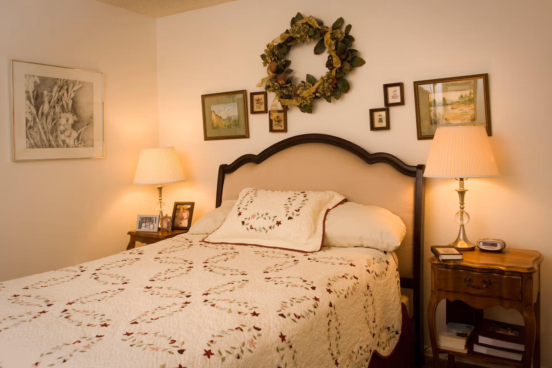 A cozy bedroom with a bed featuring a quilted bedspread and matching pillow with floral patterns. The bed has a wooden headboard, and there are two wooden nightstands on either side, each with a lamp. The walls are decorated with framed pictures and a wreath above the bed.