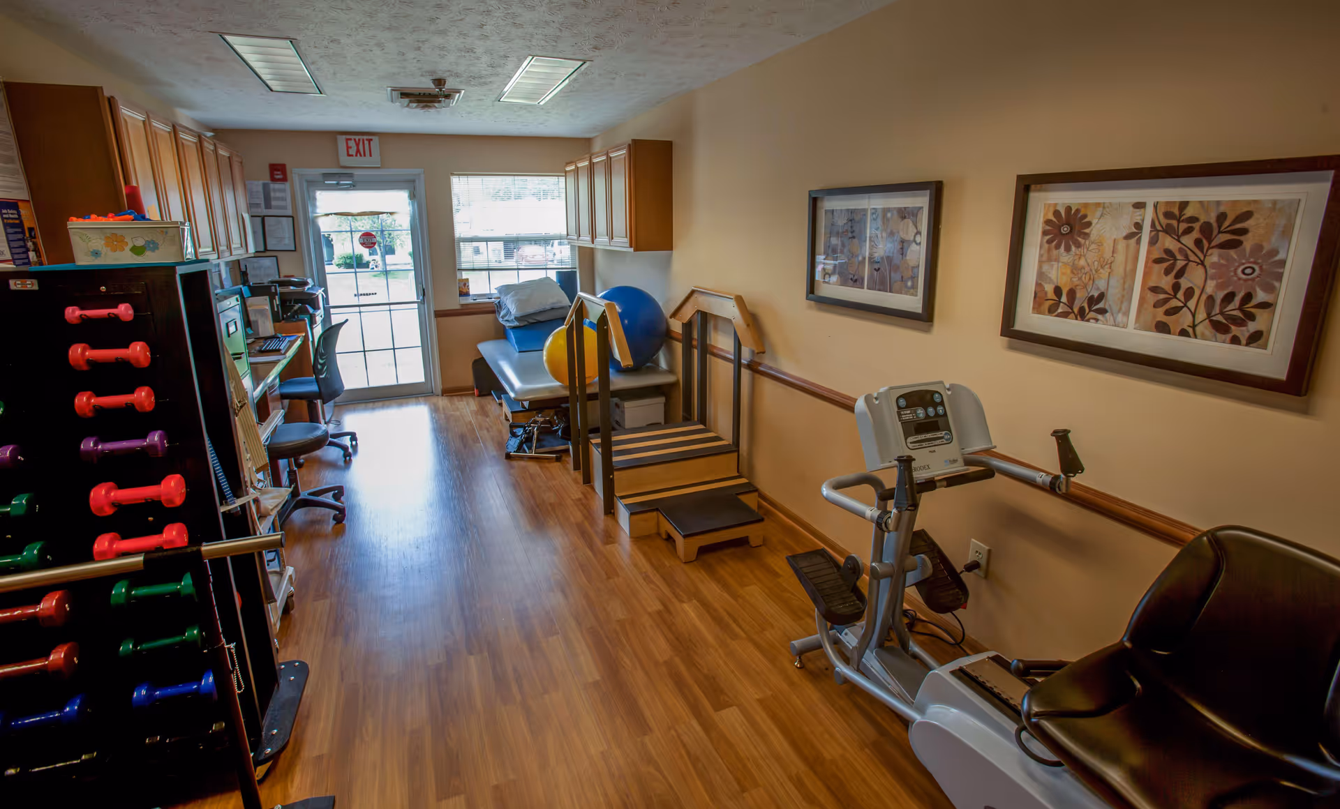 A small physical therapy or exercise room with wooden flooring, light beige walls, and ceiling lights. The room contains a rack of colorful dumbbells on the left, a computer workstation with a chair, a padded therapy table with exercise balls, a set of stairs with handrails for rehabilitation, and a recumbent exercise bike. Two framed floral artworks hang on the right wall, and a door with an exit sign and a window letting in natural light is visible at the far end.