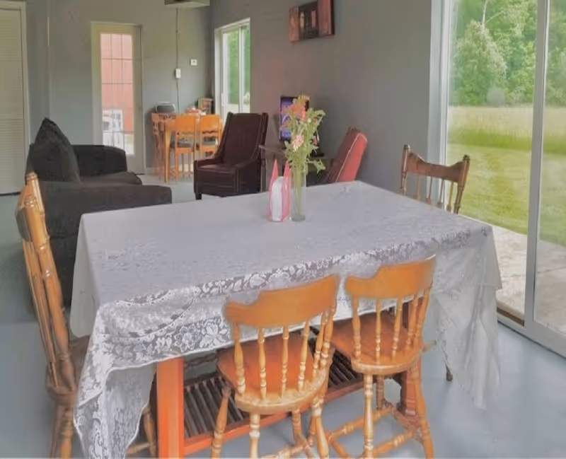 Dining table with a lace tablecloth and wooden chairs in a room opening to a living area and a sliding glass door to a grassy yard.