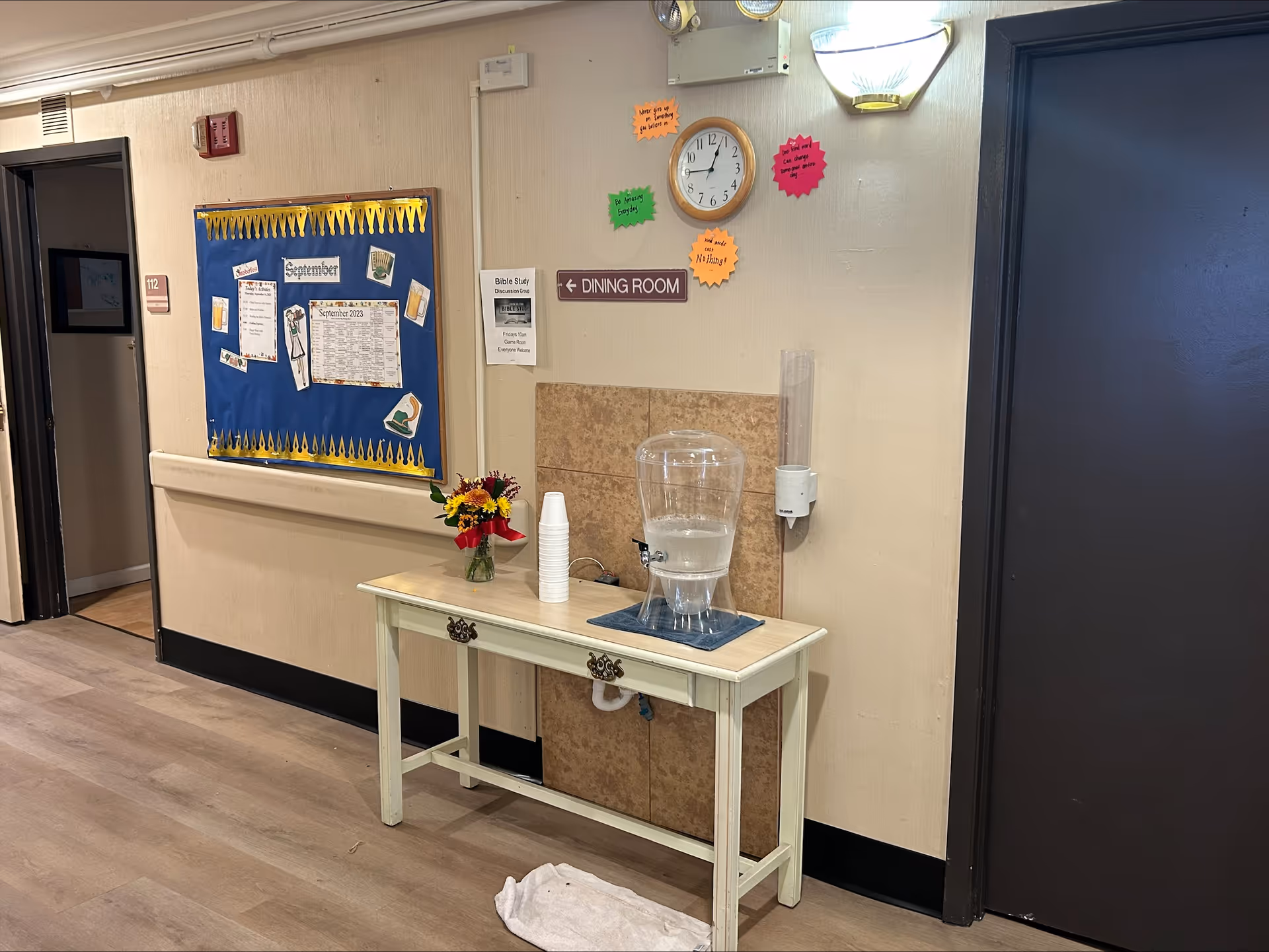 Indoor hallway area with a small table holding a water dispenser, a stack of disposable cups, and a vase with flowers. Above the table is a wall clock and a sign pointing left to the dining room. A bulletin board with various papers and a calendar is mounted on the wall to the left. There is a dark door on the right side and an open doorway on the left side of the hallway.