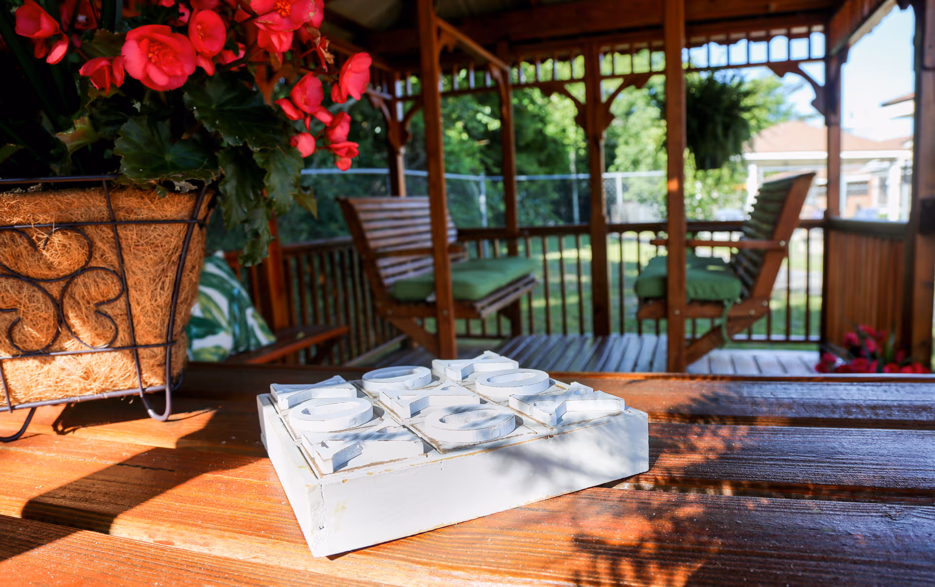 A wooden outdoor gazebo with green cushioned chairs and a wooden table. On the table, there is a white wooden tic-tac-toe game and a hanging basket with vibrant red flowers. The background shows a fenced yard with trees and a building.