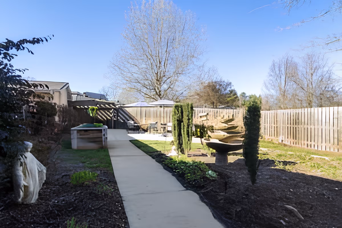 Outdoor garden area with a concrete pathway leading to a seating area with tables and umbrellas. The garden is bordered by a wooden fence and has small trees and plants along the pathway. The sky is clear and blue.