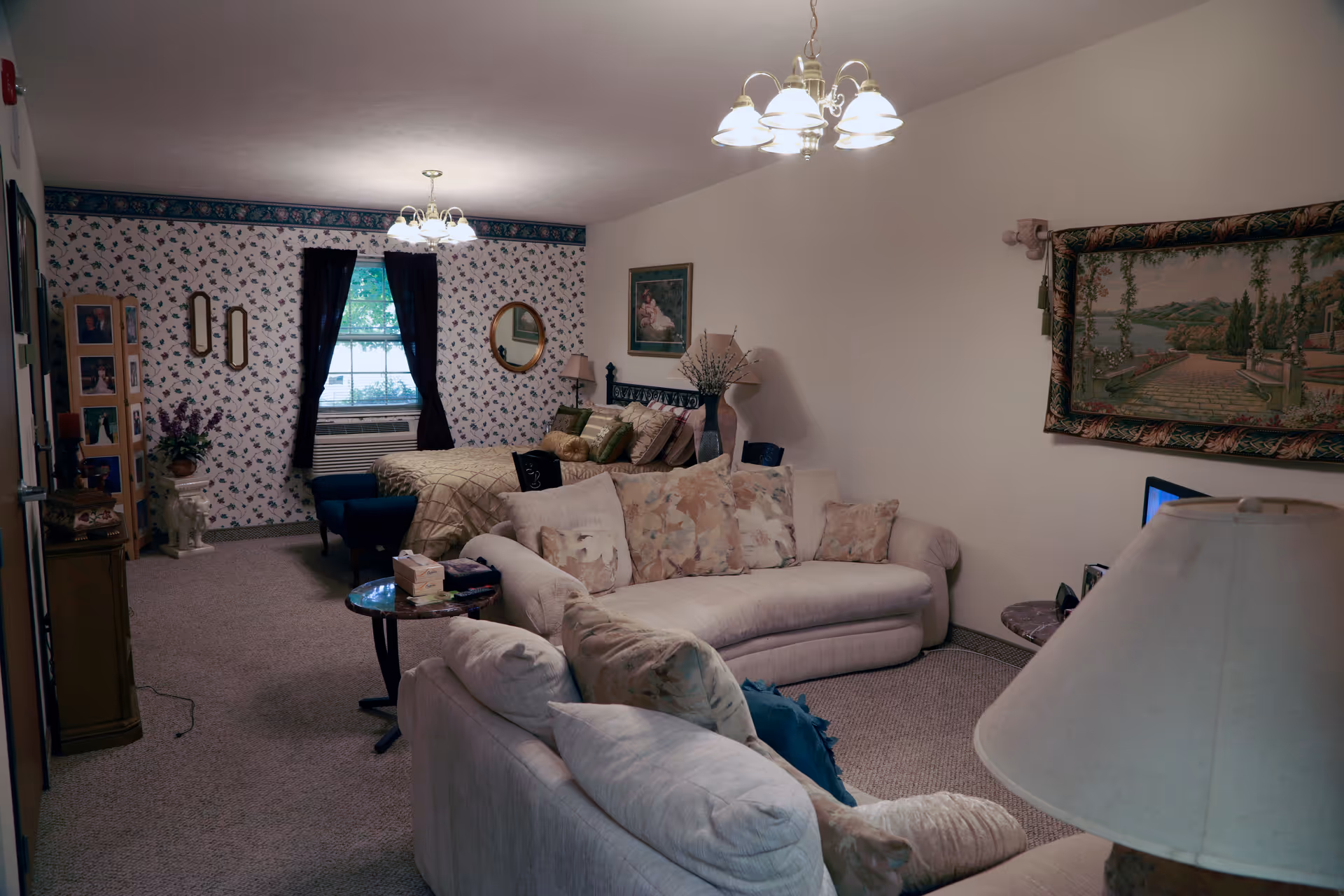 A cozy living space in an assisted living facility featuring a floral patterned wallpaper on one wall with a window covered by dark curtains. The room includes a bed with multiple pillows, a small blue ottoman, a beige sofa with floral cushions, a side table with a lamp, and framed artwork on the walls. The carpeted floor and soft lighting create a warm and inviting atmosphere.