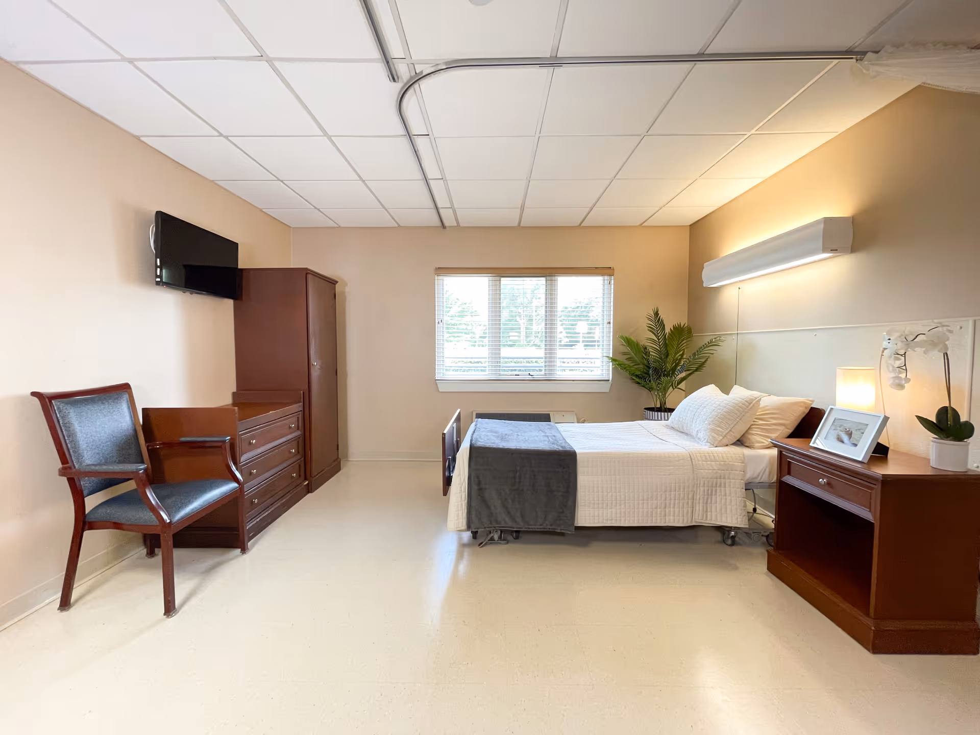 A tidy nursing facility bedroom with a single bed, bedside table, dresser, chair, wall-mounted TV and a window.
