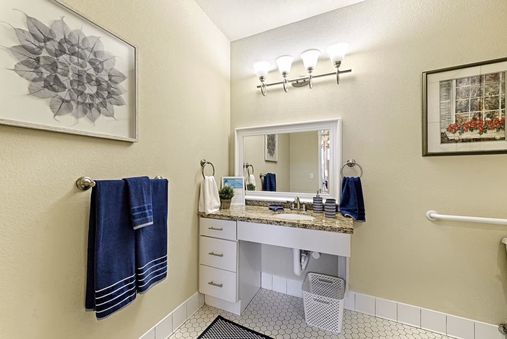 Well-lit bathroom with a granite-topped vanity, mirror, cabinets, blue towels, and framed wall art.