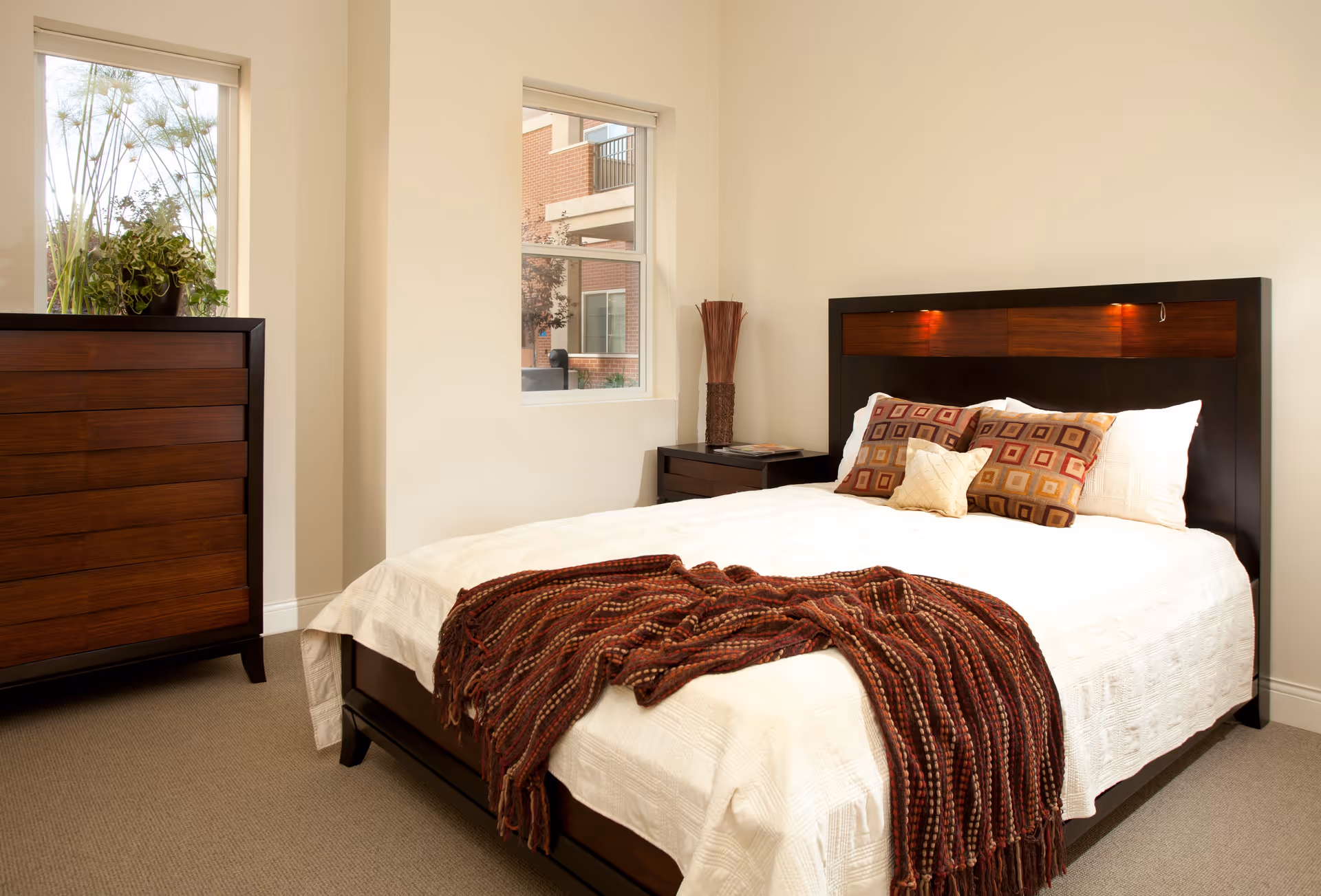 A neatly made bed with white bedding and a brown and red patterned throw blanket at the foot. The bed has a dark wooden headboard with built-in lights. There are three decorative pillows on the bed. To the left of the bed is a dark wooden dresser with a potted plant on top. A small dark wooden nightstand with a decorative vase and a magazine is next to the bed. Two windows let in natural light, showing a partial view of a brick building outside.