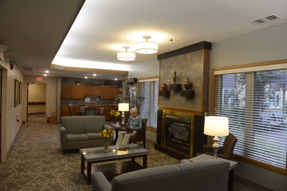 A cozy senior living facility common area with two gray sofas, a glass coffee table with a vase of yellow flowers, a fireplace with decorative vases on shelves above it, large windows with blinds, and a woman sitting in a chair reading a book. The background shows a dining area with wooden cabinets and chairs.