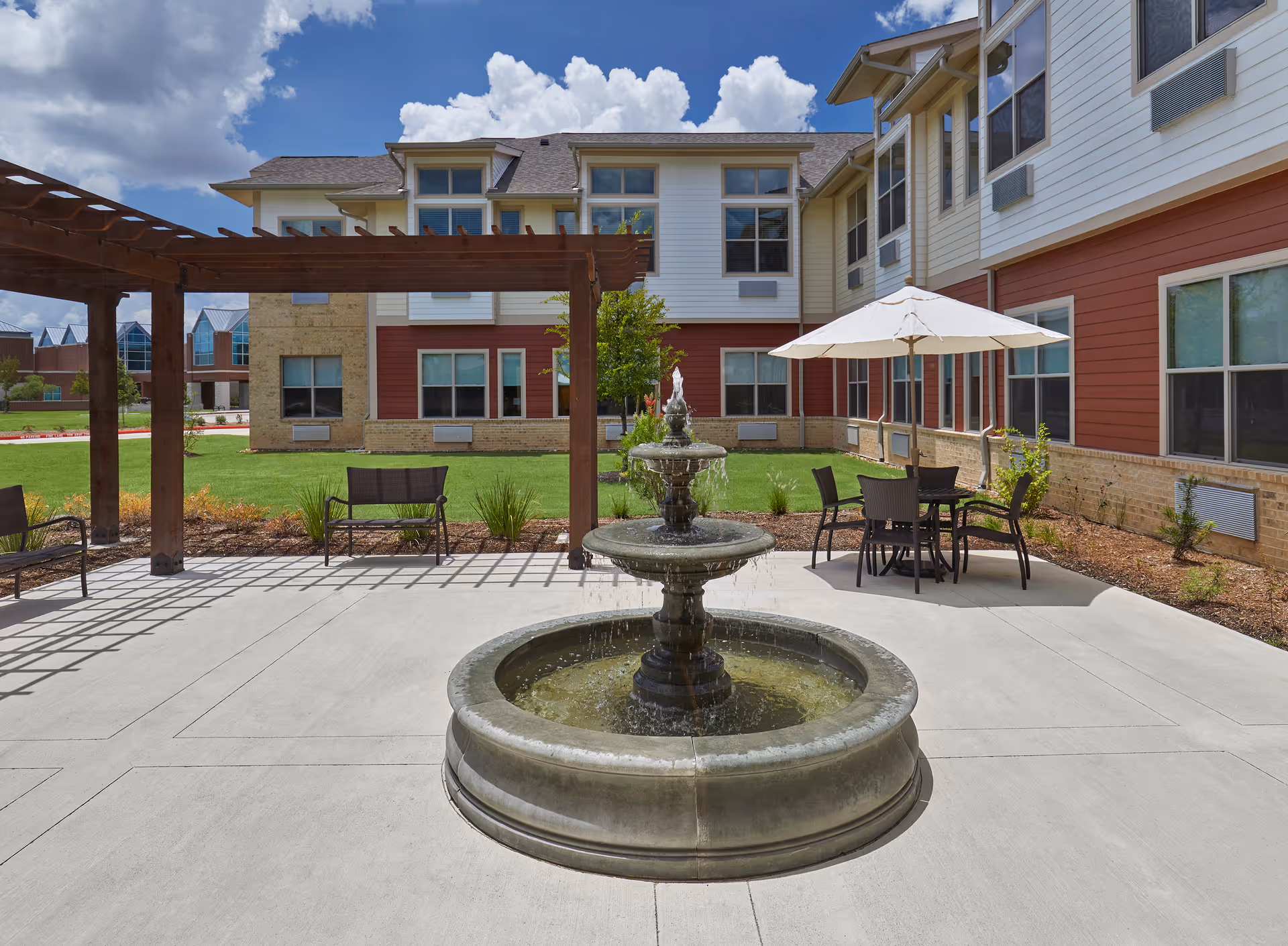 Outdoor courtyard area of a senior living facility with a three-tiered water fountain in the center, surrounded by concrete pavement. There is a wooden pergola with benches underneath on the left side, and a round table with four chairs and a white umbrella on the right side. The building has multiple windows and a combination of red and beige siding with brick accents. The sky is partly cloudy.