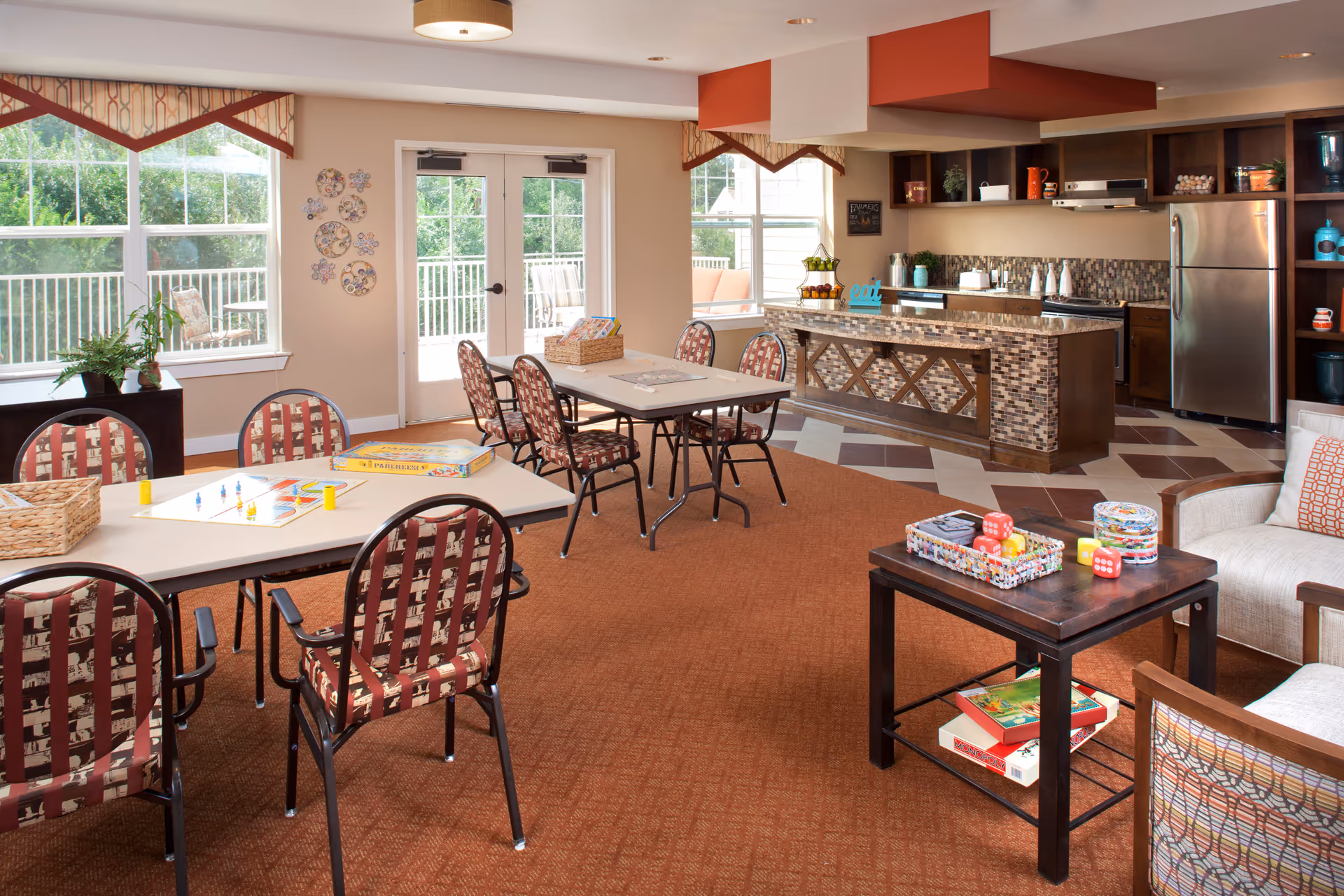 A bright and spacious common area in an assisted living facility featuring multiple tables and chairs arranged for games and activities. The room has large windows and glass doors letting in natural light, with a view of an outdoor patio. There is a kitchen area with a mosaic tile island, stainless steel refrigerator, and open shelving. The room is decorated with wall art, plants, and board games on the tables and coffee table.