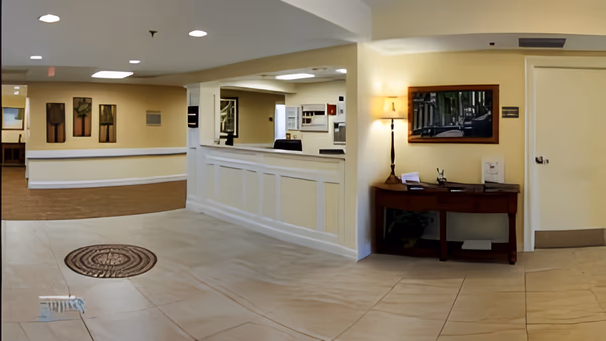 Interior view of a senior living facility reception area with a white reception desk, beige walls, and tiled floor. A wooden table with a lamp and some papers is placed against the wall, and a framed picture hangs above it. The area is well-lit with ceiling lights.