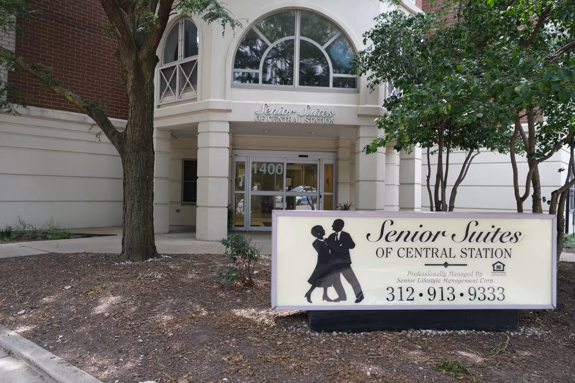 Front entrance of Senior Suites of Central Station with a sidewalk, trees, and a large sign displaying the name and phone number.