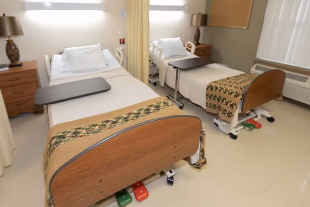 Two adjacent adjustable hospital-style beds in a shared patient room with overbed tables and bedside furniture.