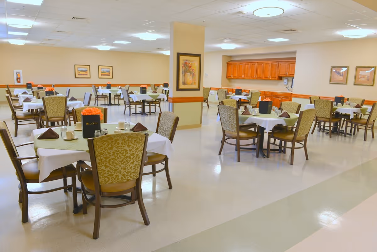A spacious dining room in a senior living facility with multiple tables covered with white and green tablecloths, each set with cups, napkins, and menus. The room has beige walls, framed artwork, and wooden cabinetry along one wall. The floor is light-colored and the ceiling has recessed lighting.