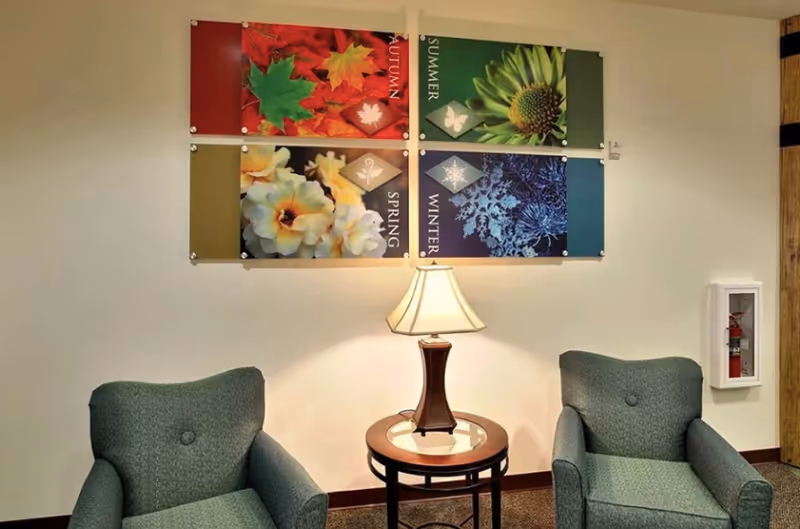 Two upholstered armchairs flank a round side table with a lamp beneath colorful seasonal wall art in a lounge area.