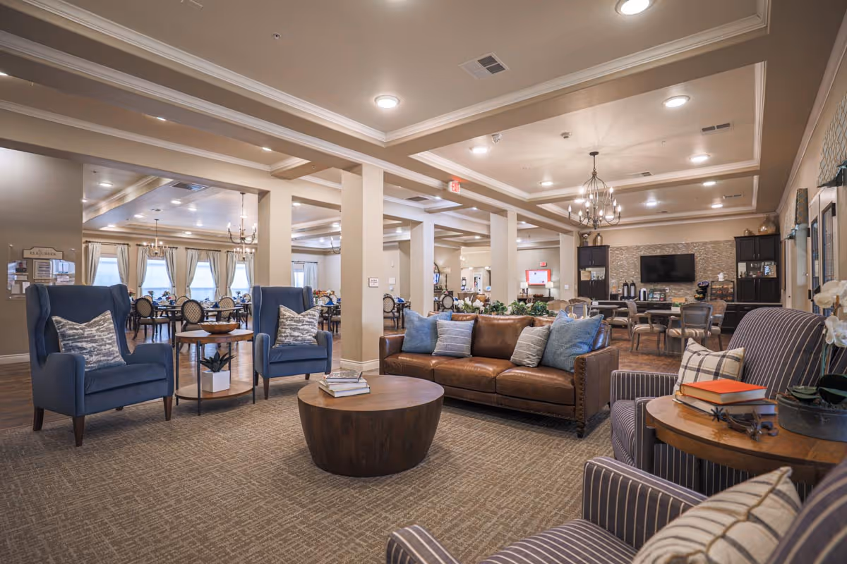 A spacious and well-lit common area in an assisted living facility featuring comfortable seating including a brown leather sofa, blue armchairs, and striped chairs with pillows. There is a round wooden coffee table with books on it, and a side table with more books and a plant. In the background, there are dining tables and chairs, chandeliers, and a wall-mounted TV with cabinets below it. The room has beige walls, carpeted flooring, and large windows with curtains.