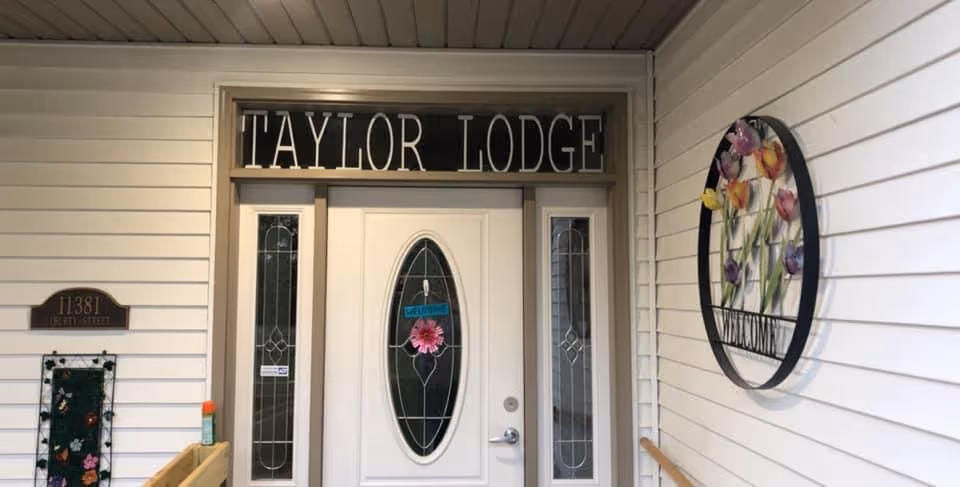 Entrance door of Taylor Lodge with a white door featuring an oval glass panel decorated with a pink flower and a small welcome sign. The door is flanked by two narrow windows with decorative glass. Above the door is a sign that reads 'TAYLOR LODGE'. To the left of the door is a plaque with the address '11381' and a small decorative item below it. On the right wall, there is a round floral wall decoration with the word 'WELCOME'. The exterior walls are covered with white siding.