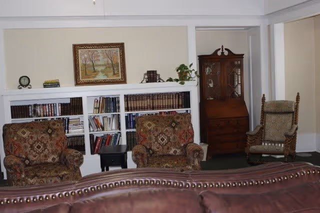 A cozy living room area with two patterned armchairs and a wooden chair with cushions. Behind the chairs is a built-in white bookshelf filled with books and decorative items, and above it hangs a framed painting of a landscape with trees. To the right is a wooden cabinet with glass doors and a small plant on top. The foreground shows the back of a brown leather sofa.