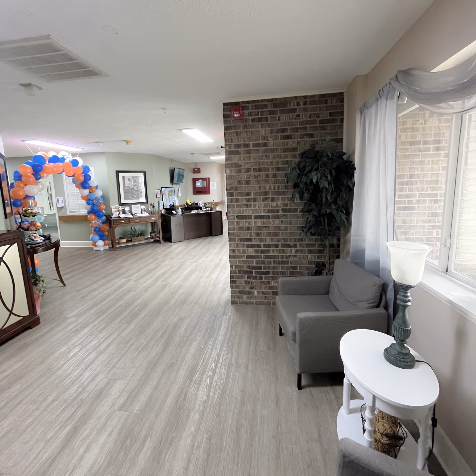 Interior view of a senior living facility hallway with light wood flooring, a gray armchair next to a white side table with a lamp, a large window with sheer curtains, a brick accent wall, and a reception desk area decorated with a colorful balloon arch in the background.
