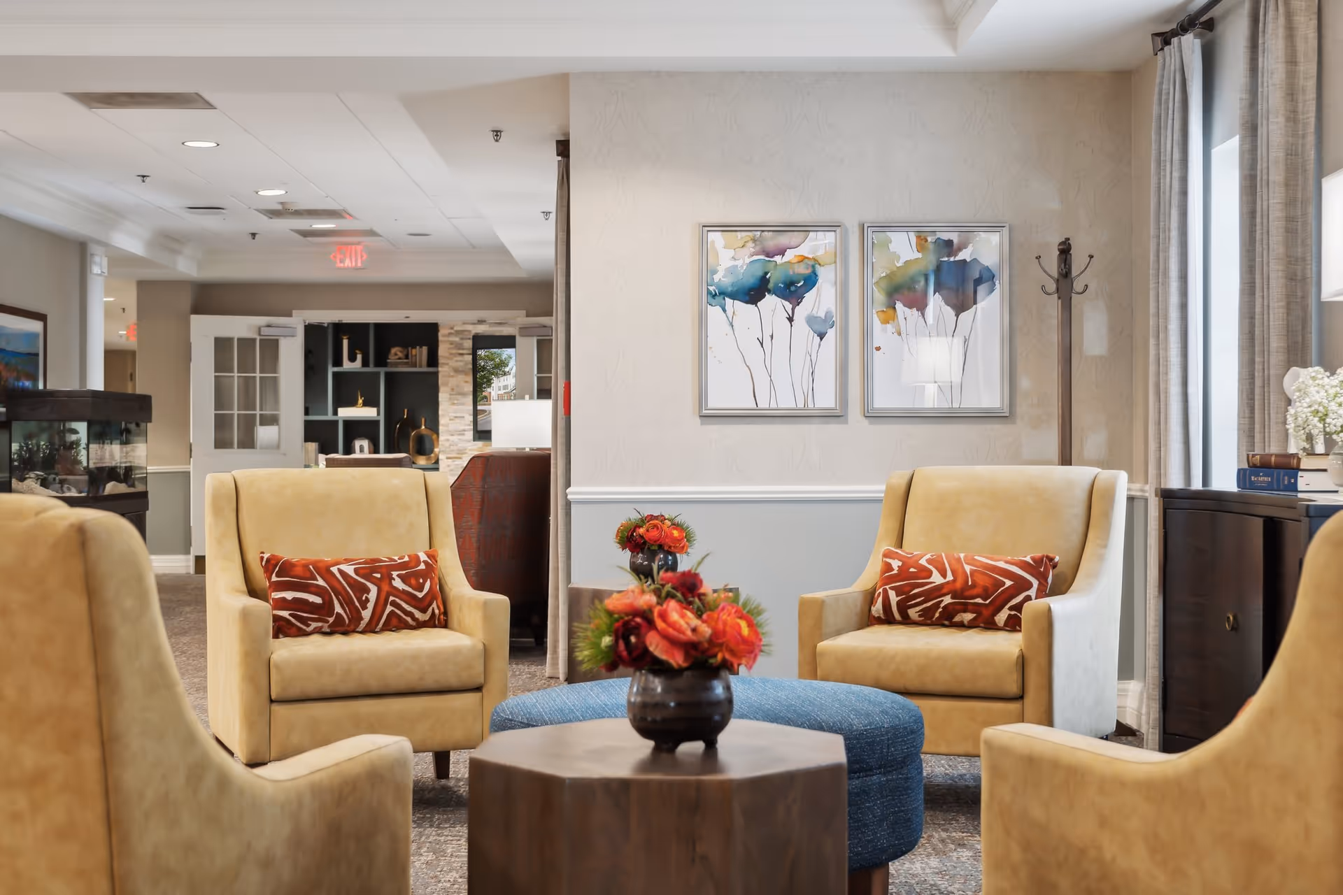A bright lounge with beige armchairs arranged around a central table with a flower arrangement, a blue ottoman, and framed artwork on the wall.