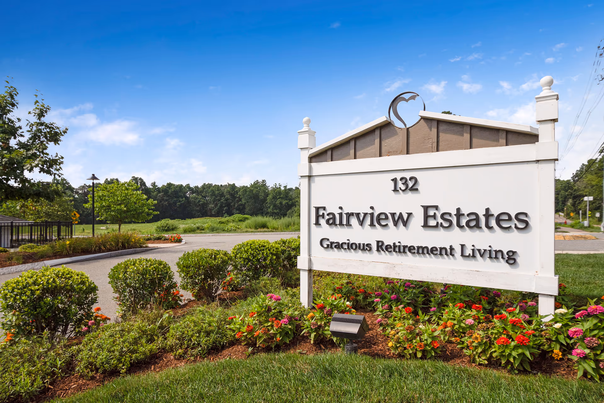 Outdoor view of the entrance sign for Fairview Estates Gracious Retirement Living, surrounded by landscaped bushes and colorful flowers under a clear blue sky.