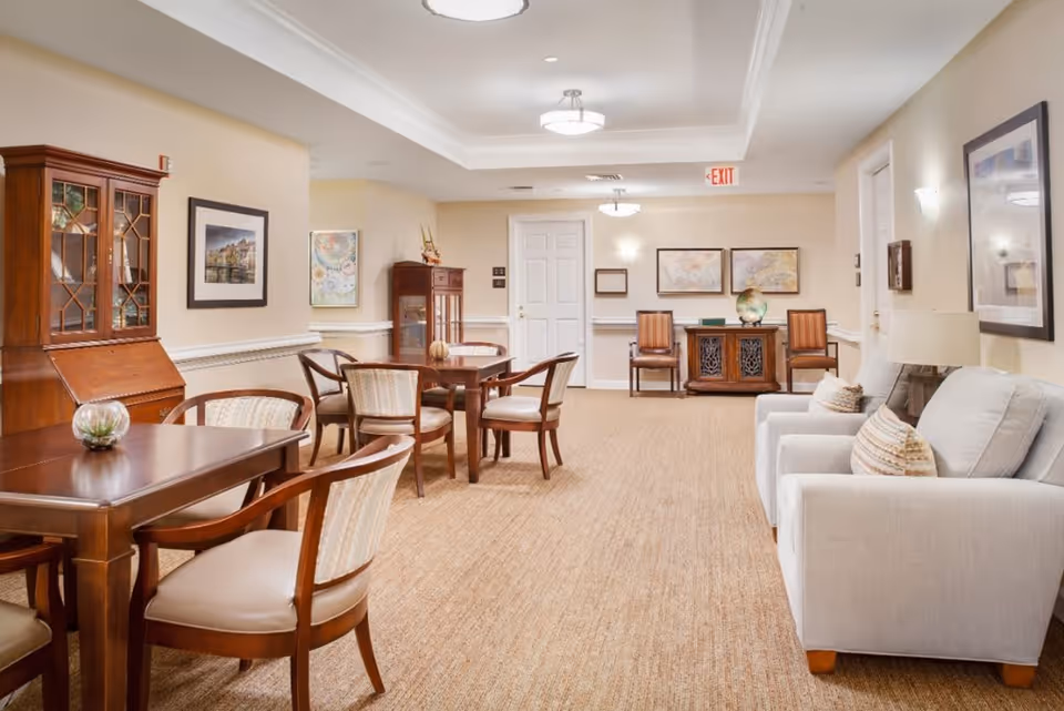 A well-lit common area in a senior living facility featuring beige walls and carpet. The room includes wooden tables with upholstered chairs, a light gray sofa with patterned pillows, framed artwork on the walls, and decorative cabinets. Ceiling lights and wall sconces provide illumination, and an exit sign is visible above a white door at the back.