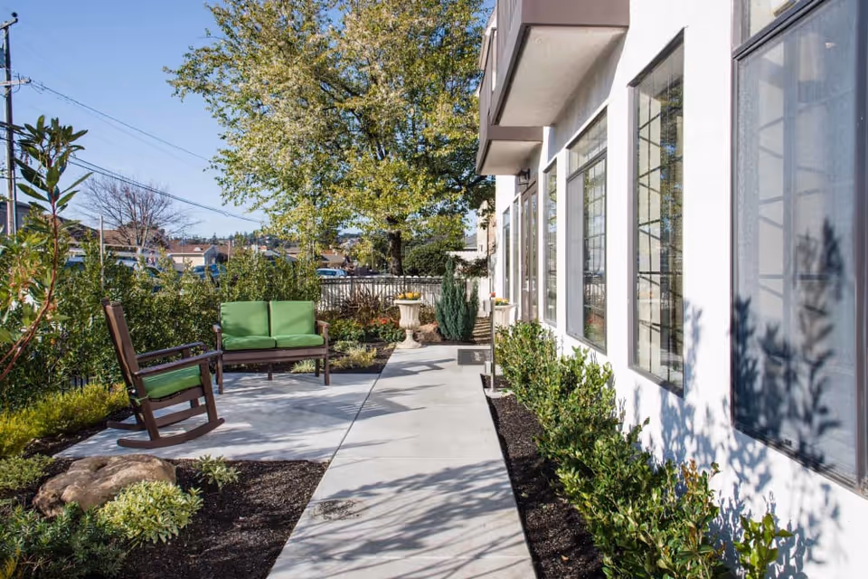 Outdoor patio area at Carefield Castro Valley Assisted Living & Memory Care featuring a concrete walkway, green cushioned wooden bench and rocking chair, surrounded by plants and shrubs with a large tree in the background under a clear blue sky.