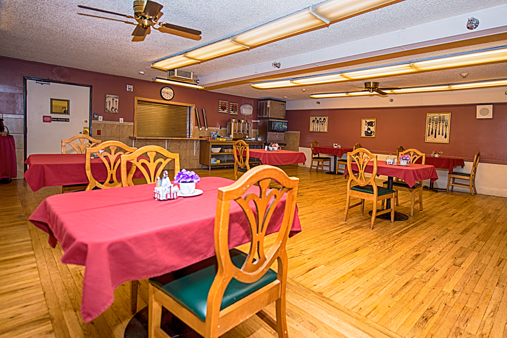 Spacious dining room with wooden chairs and tables covered in red tablecloths and ceiling fans.