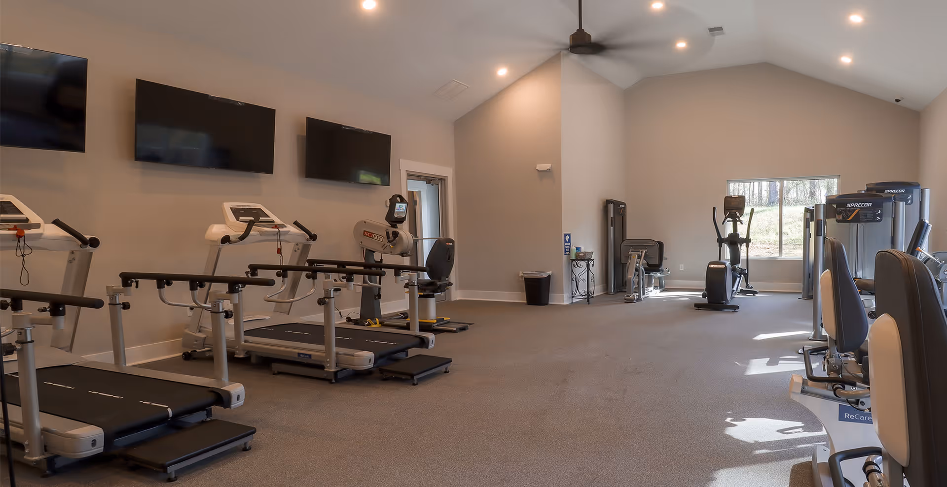 A spacious fitness room with several treadmills, exercise bikes, and strength training machines. The room has beige walls, a high ceiling with recessed lighting and a ceiling fan, and a large window letting in natural light. Three flat-screen TVs are mounted on the wall above the treadmills.
