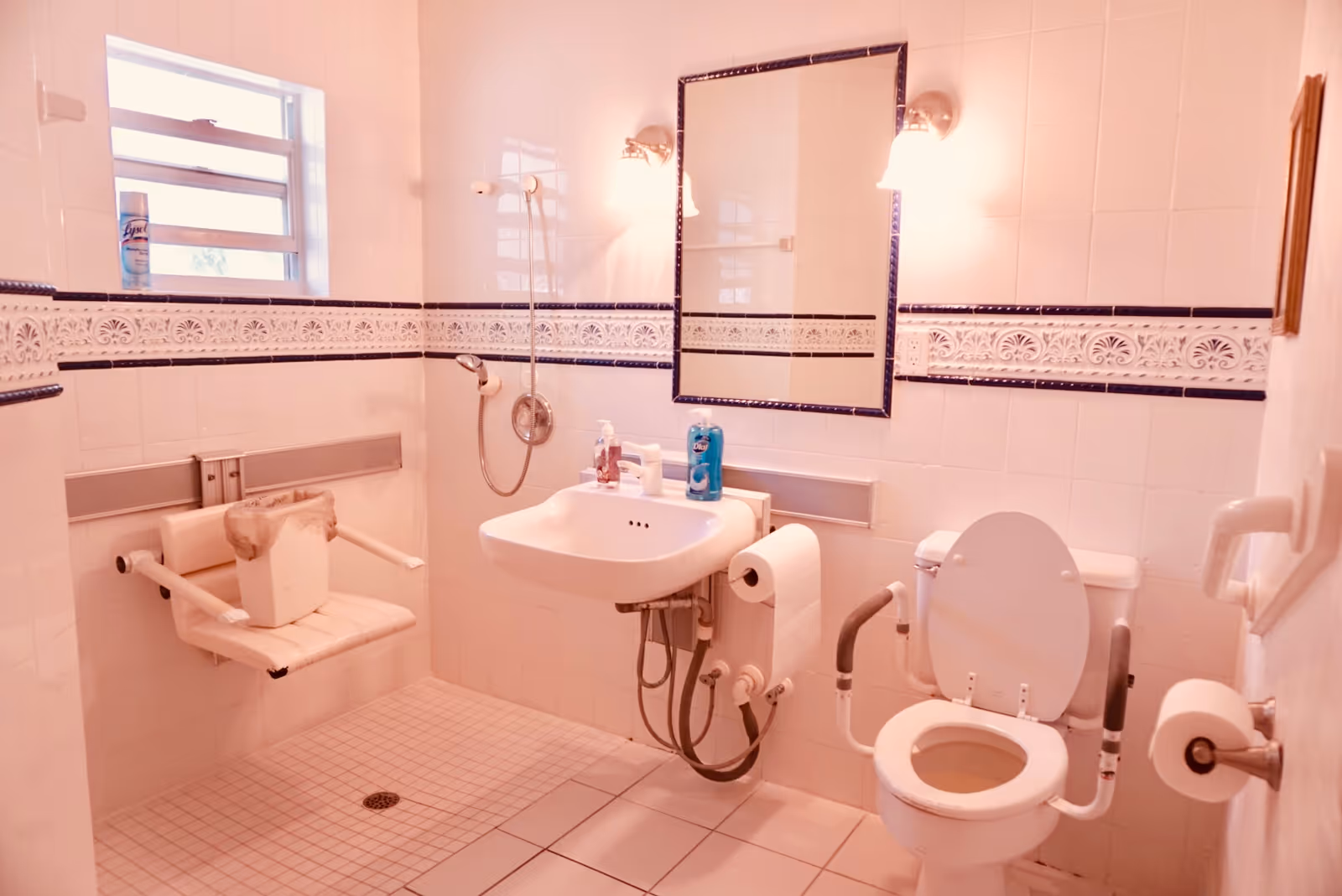 Accessible tiled bathroom with a wall-mounted sink, mirror, grab-bar-equipped toilet, handheld showerhead and a fold-down shower seat.