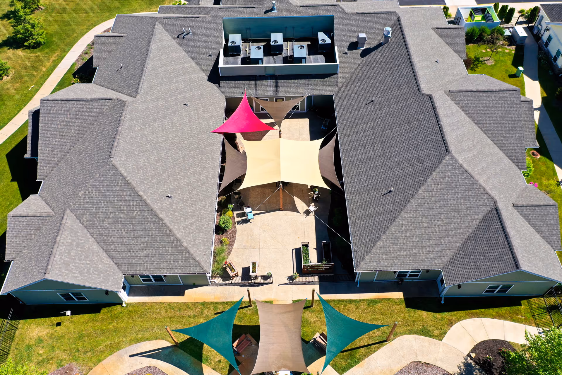 Aerial view of a large building with a central courtyard featuring colorful shade sails in red, beige, and brown. The building has a gray shingled roof and is surrounded by green lawns and paved walkways.