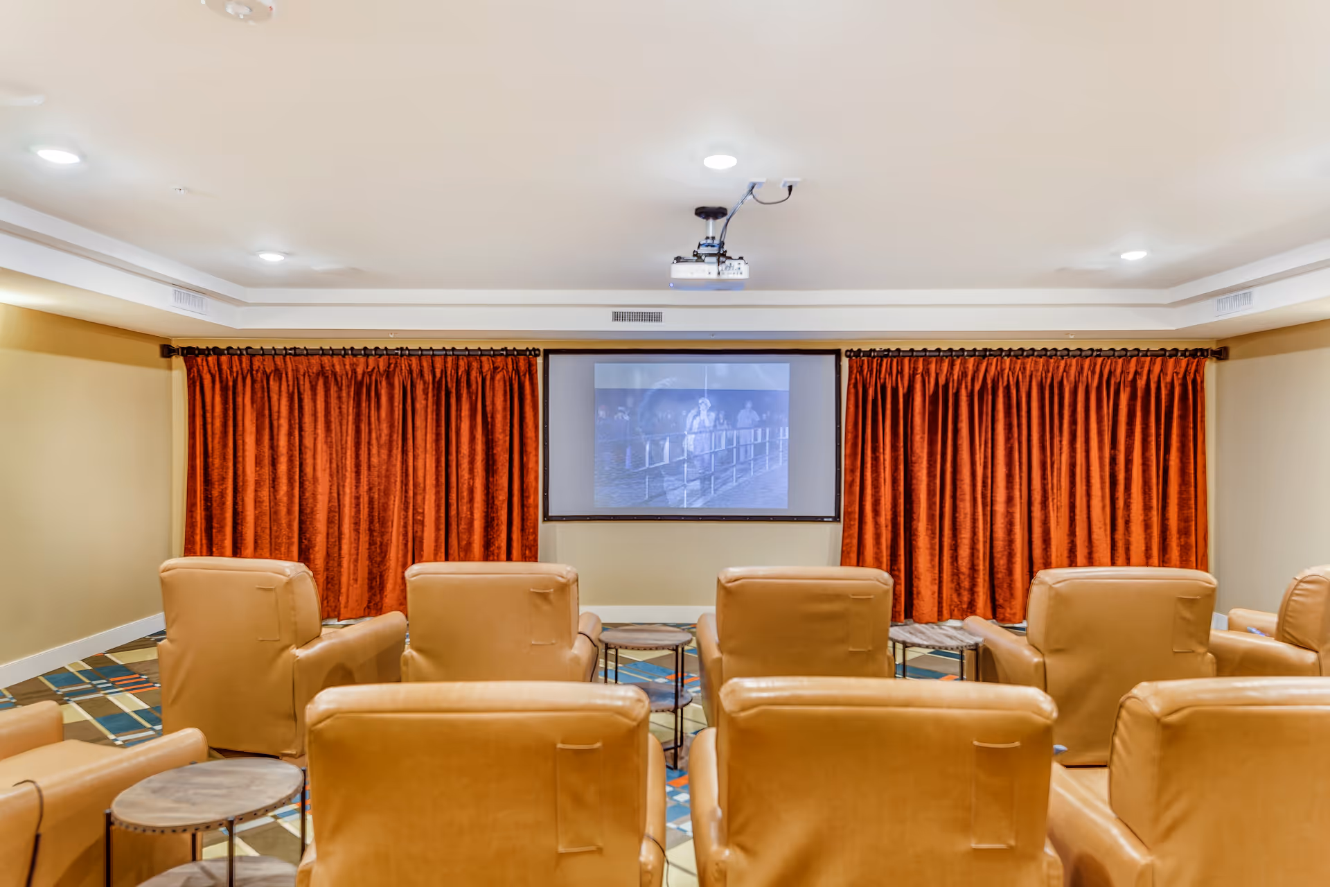 Small theater-style media room with rows of tan leather recliners facing a wall-mounted projection screen and red curtains.