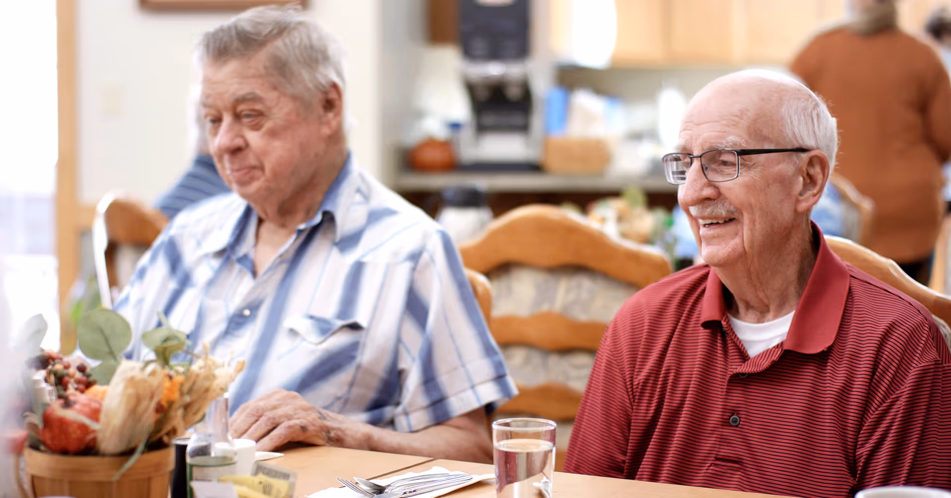 Two elderly men sitting at a dining table in a communal dining area, one wearing a red polo shirt and glasses smiling, the other in a blue and white striped shirt looking down. The table has a glass of water, utensils, and a decorative basket with autumn-themed items.