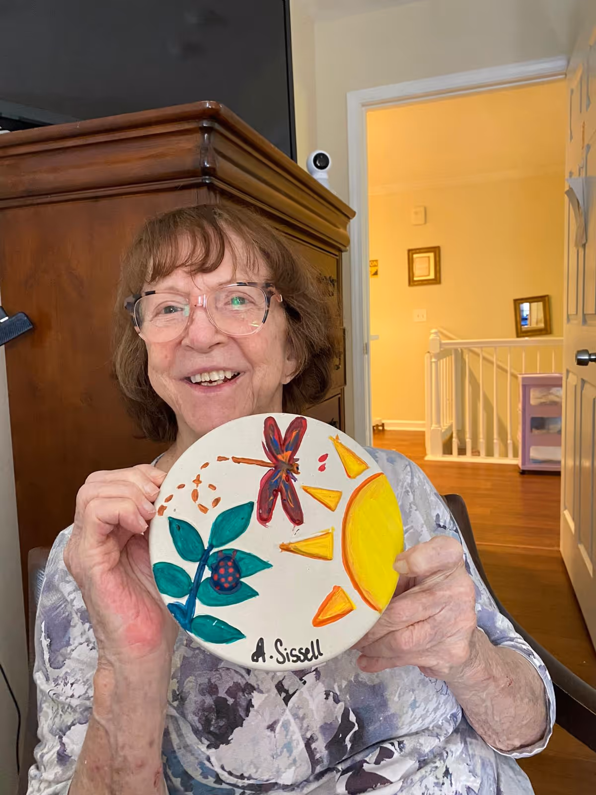 An elderly woman with glasses smiling and holding a round painted ceramic plate featuring a sun, a dragonfly, and green leaves. She is seated indoors with a wooden cabinet and an open doorway in the background.