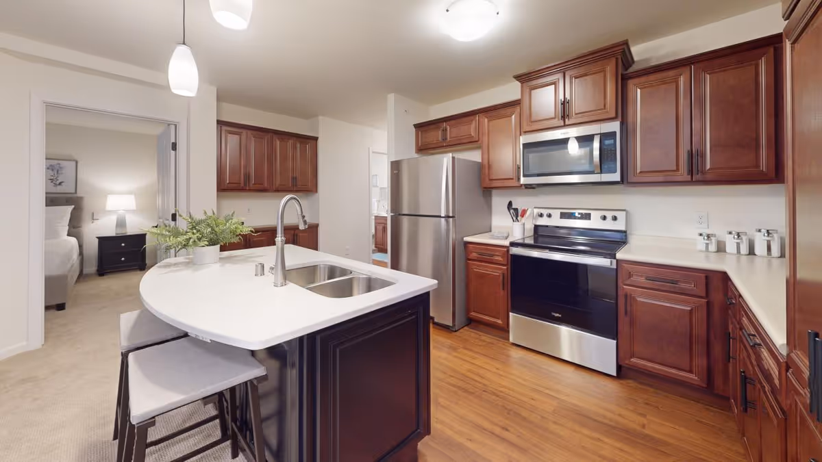 Modern kitchen with wooden cabinets, stainless steel refrigerator, oven, and microwave. A white countertop island with a sink and two stools is in the foreground. A doorway leads to a bedroom with a bed, nightstand, and lamp visible.