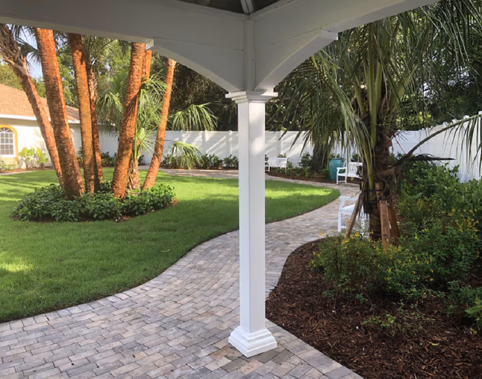 A landscaped outdoor garden area with a curved paved walkway, green grass, palm trees, bushes, and white benches. The scene is viewed from under a white gazebo structure.