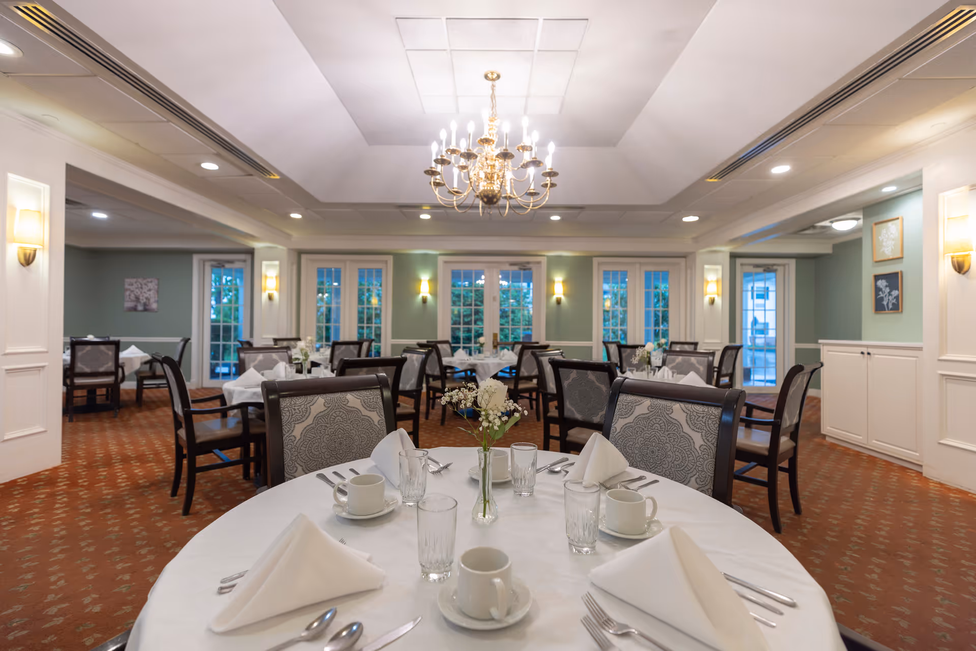 Upscale dining room with round tables set for a meal, upholstered chairs, a central chandelier, and windows along the back wall.