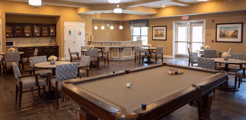 A spacious common area in a senior living facility featuring a pool table in the foreground, several round tables with chairs arranged around the room, a small bar or serving counter in the back, and cabinets along the left wall. The room has warm beige walls, wood flooring, and large windows near the entrance doors letting in natural light. Decorative framed pictures hang on the walls.