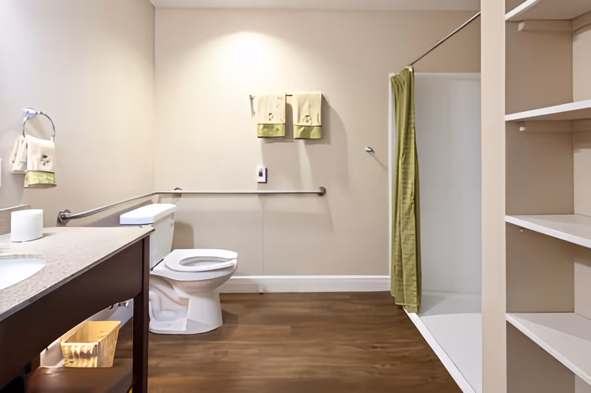 A clean and spacious bathroom with a toilet, a sink with a countertop, a shower area with a green curtain, and open shelving. The walls are light-colored, and there are two towels hanging on the wall above a grab bar. The floor is wooden.