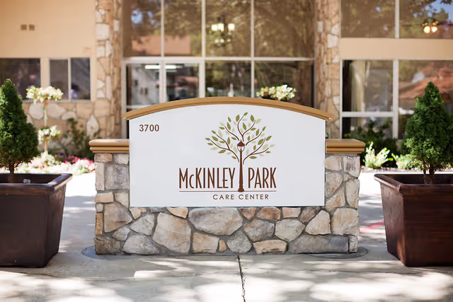 Stone sign with the text 'McKinley Park Care Center' and the number 3700, featuring a tree logo with a lamp post in the center, placed in front of a building entrance with large windows and potted plants on either side.