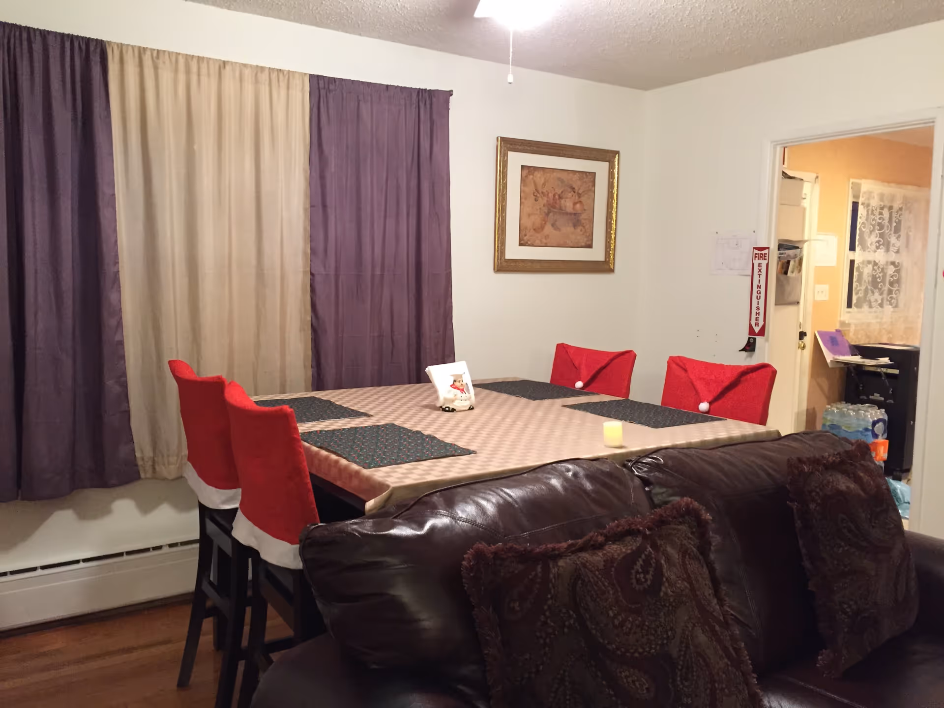 Dining area with a table covered by a tablecloth and red-covered chairs, a brown leather sofa in the foreground, and curtains on the left wall.
