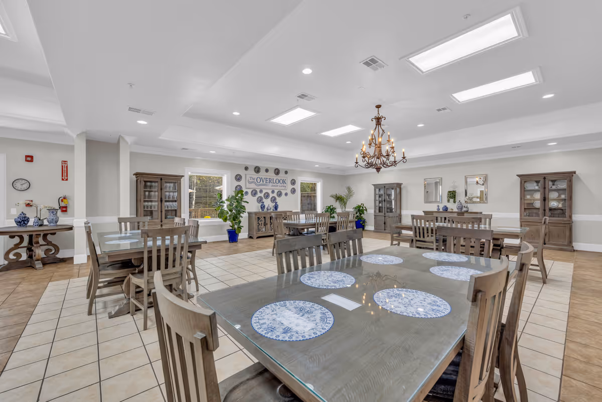 A spacious dining room with several wooden tables and chairs arranged neatly. Each table has blue and white decorative placemats. The room features a chandelier hanging from the ceiling, large windows letting in natural light, and wooden cabinets along the walls. There are also some green plants and decorative plates on the walls, including a sign that reads 'The Overlook'.