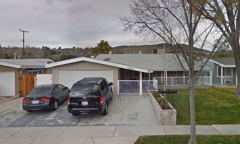 Front view of a single-story senior facility with a driveway, parked cars, and a lawn.