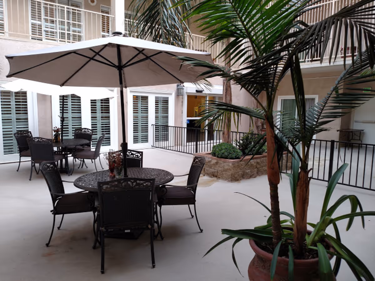 Outdoor courtyard area with round metal tables and chairs, large umbrellas providing shade, potted plants including a tall palm, and a building with multiple windows and balconies surrounding the space.