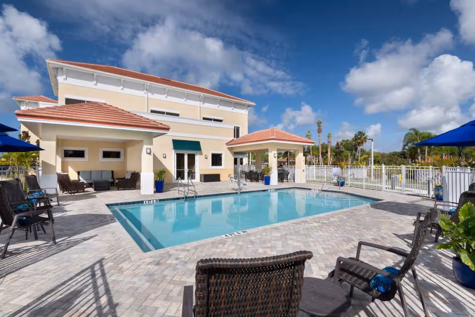 Outdoor swimming pool and patio with lounge chairs and umbrellas in front of a two-story building under a blue sky.