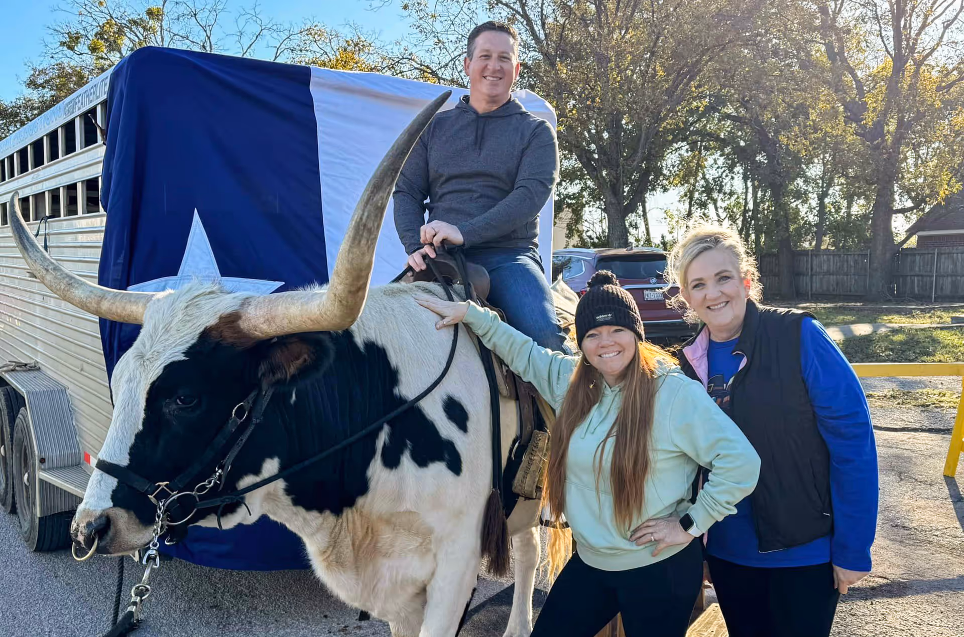 A man is sitting on a large black and white longhorn steer with prominent horns. Two women stand beside the steer, smiling at the camera. Behind them is a trailer with a large Texas flag draped over it, and trees and parked cars are visible in the background.