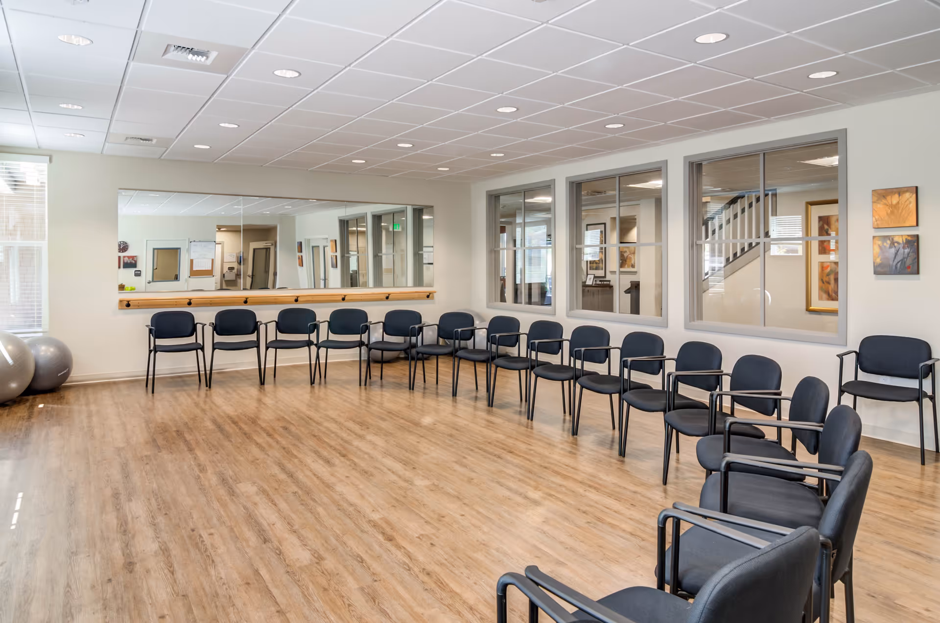 A spacious room with wooden flooring and a large mirror on one wall. There are multiple black chairs arranged in a semi-circle around the room. Three exercise balls are placed in the corner near a window with blinds. The room has a white ceiling with recessed lighting and several windows looking into an adjacent area with framed artwork on the walls.