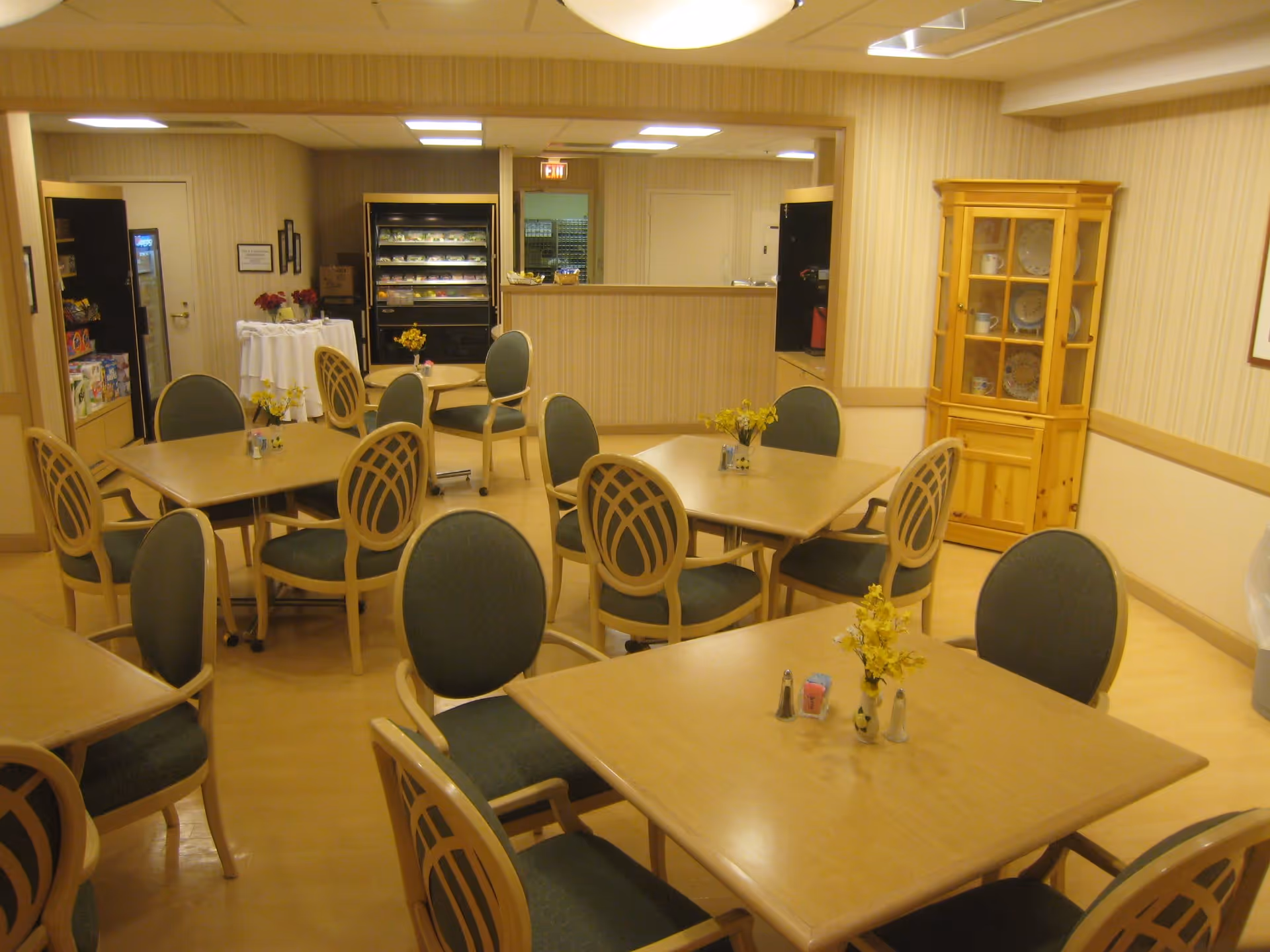 A dining room with multiple square wooden tables and green cushioned chairs arranged around them. Each table has a small vase with yellow flowers and salt and pepper shakers. In the background, there is a counter area with a refrigerated display case and a wooden cabinet with dishes inside. The walls are light-colored with vertical stripes, and the floor is a light wood tone.