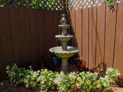 A three-tier stone fountain sits in a small garden courtyard with a wooden fence.