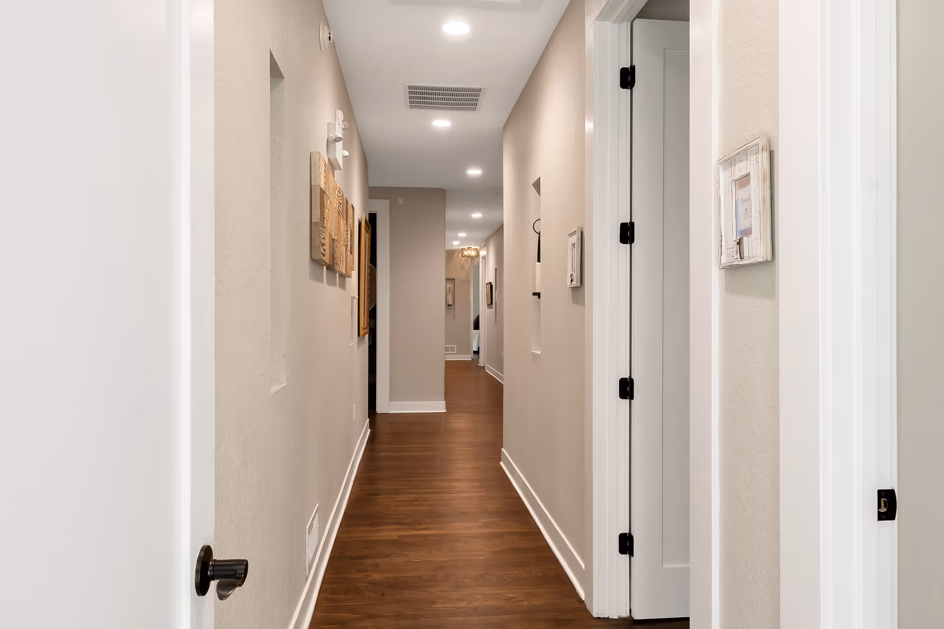 A long, well-lit hallway with wooden flooring and beige walls. Several white doors are visible on the right side, and wall decorations are hung on the left wall. Recessed ceiling lights illuminate the corridor.